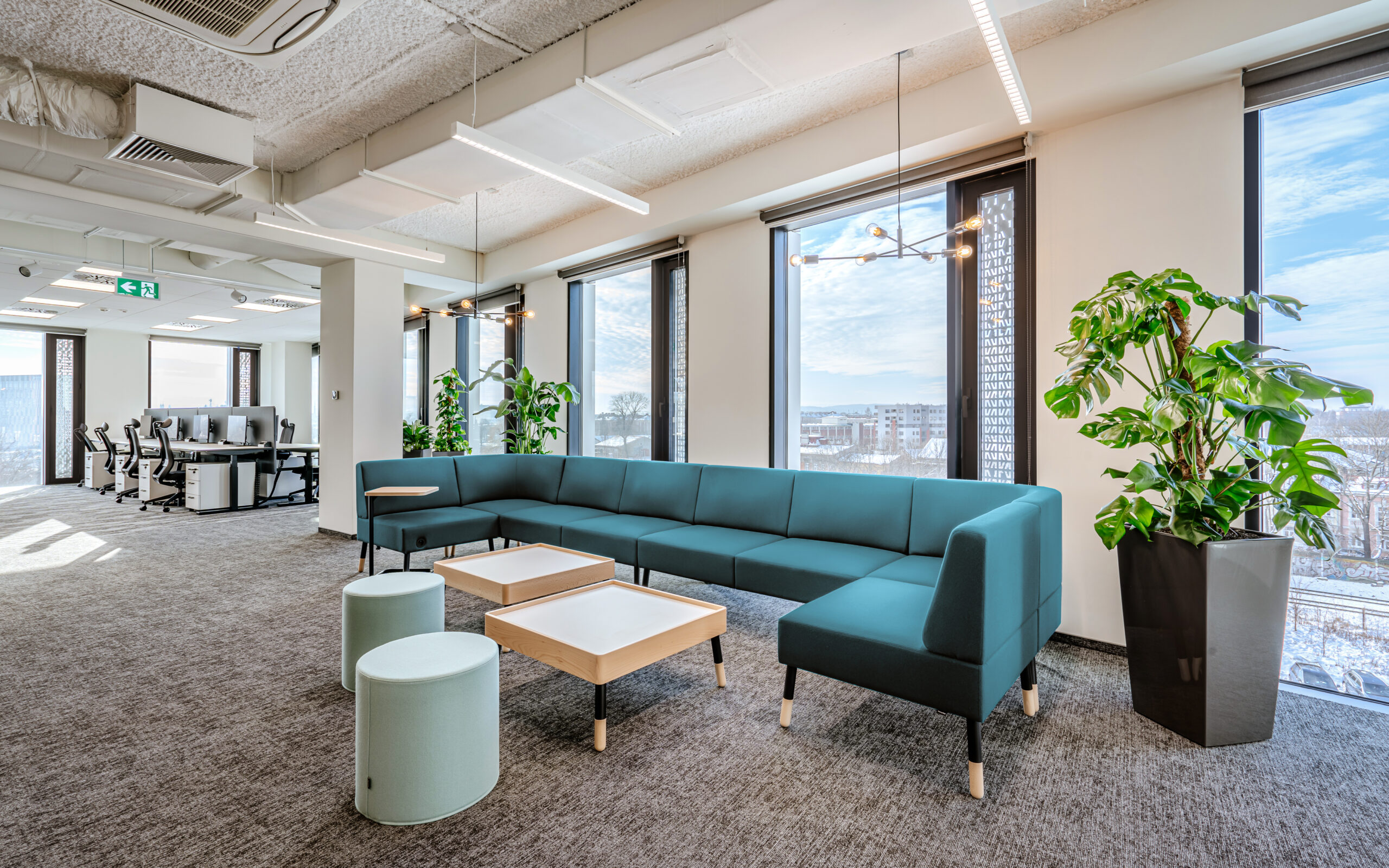 Bright and modern Krakow, Poland office space featuring a large teal sectional sofa with light wooden tables, soft ottoman seating, and lush green potted plants. Floor-to-ceiling windows offer views of the cityscape under a blue sky, with a backdrop of workstations and ergonomic chairs in an open-concept layout.