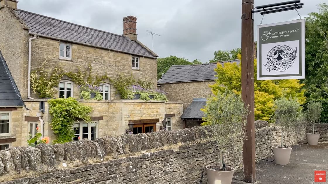A stone country inn with ivy and flowers on the facade, and a sign reading “The Feathered Nest Country Inn” in the Cotswolds.