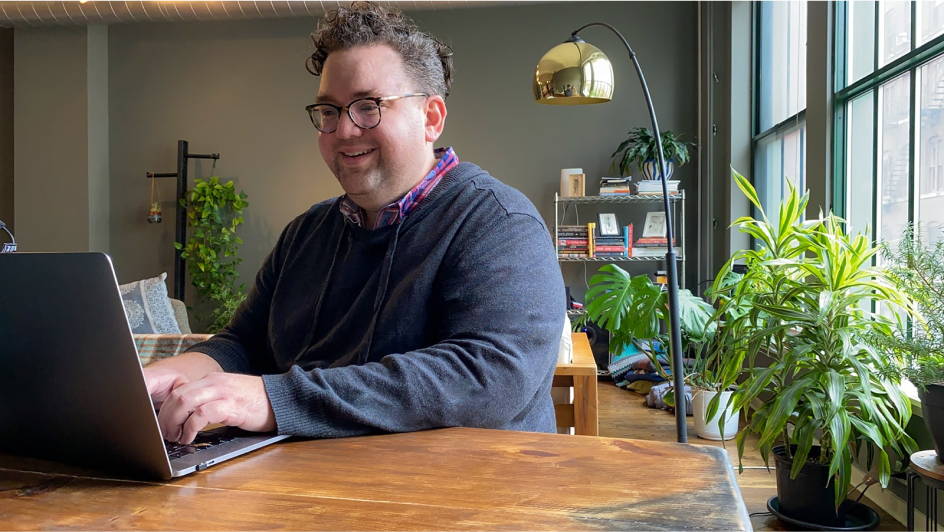 A male-identifying engineer smiles in his home office while working on his laptop.