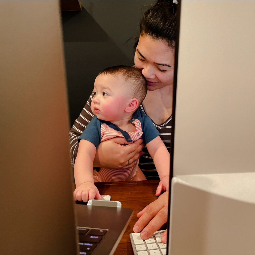 A female-identifying Sprout employee holding her small child while working on her laptop.