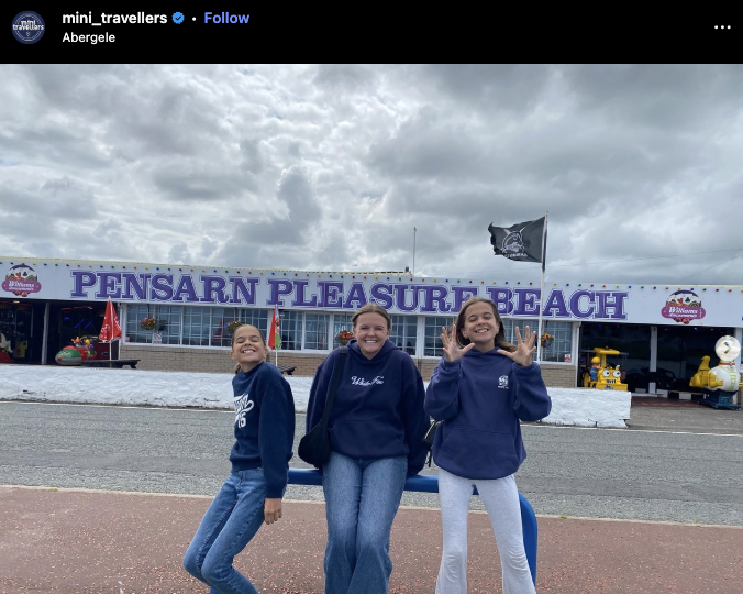 A smiling mother and two daughters pose in front of Pensarn Pleasure Beach in Abergele, Wales, wearing matching navy sweatshirts.