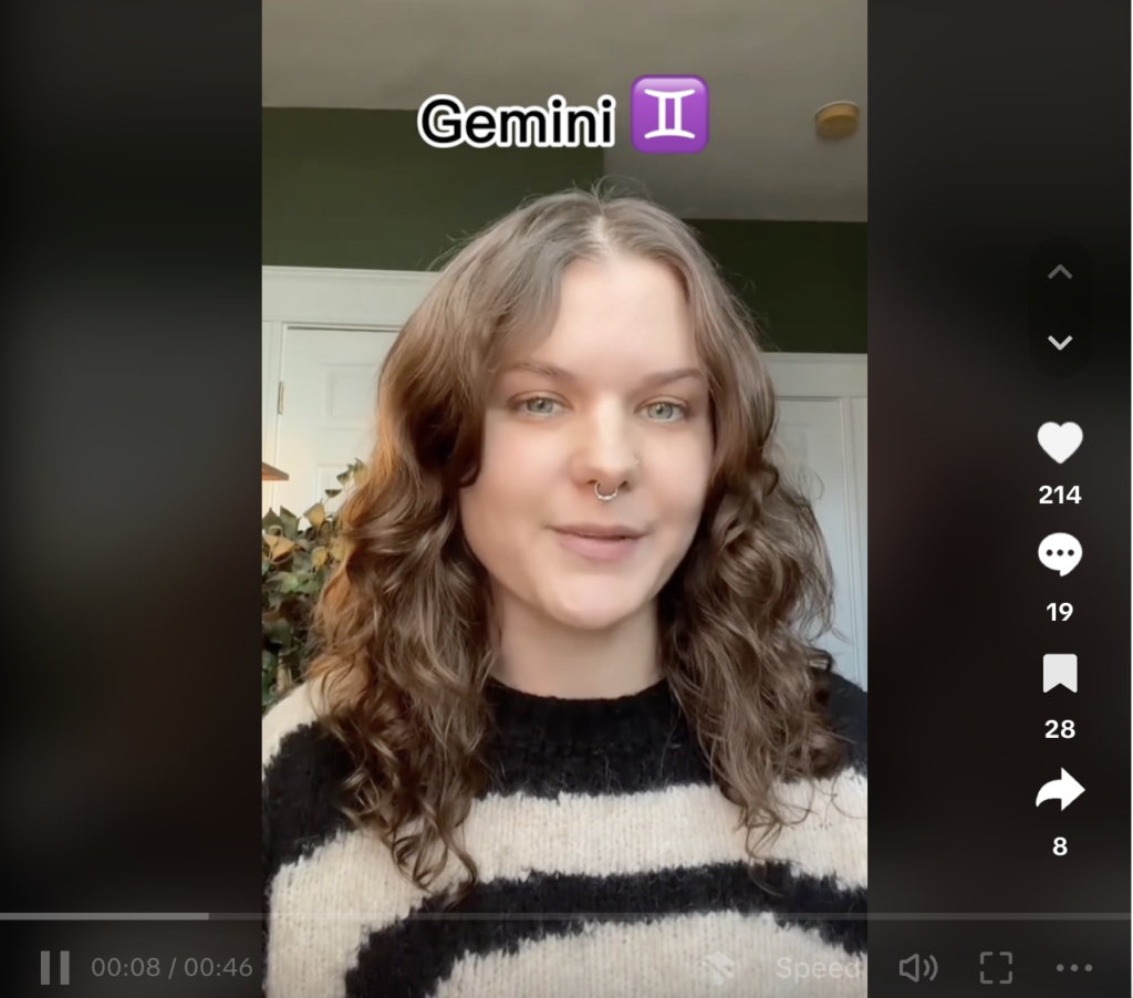 Video from Domino’s TikTok account, featuring a woman with brown hair and a septum nose ring in a striped sweater, and text on screen that reads “Domino’s Astrology Part 1.” 